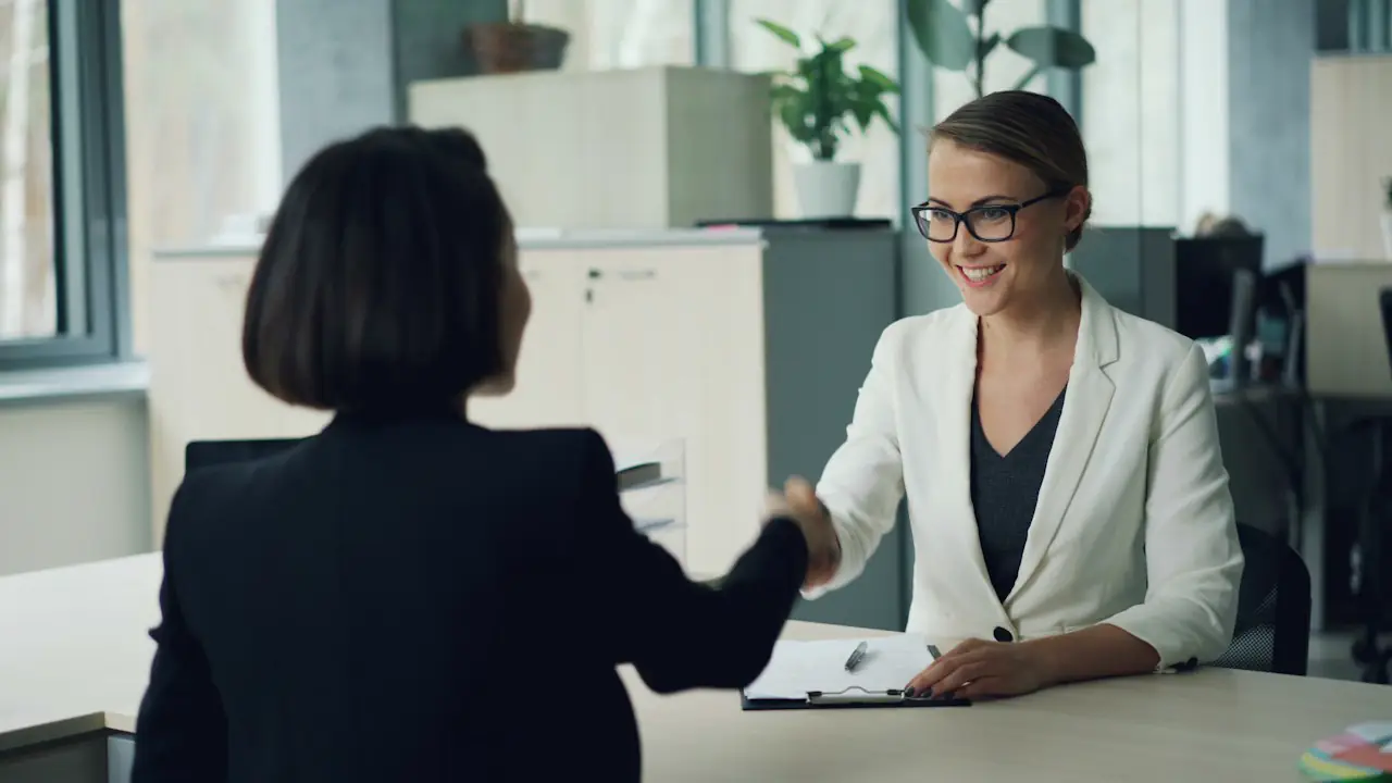 Two women shaking hands across a desk in a bright office