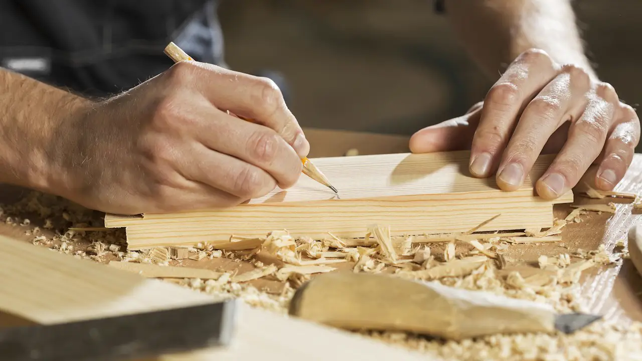 A woodworker marking a board with a pencil on a bench covered in shavings
