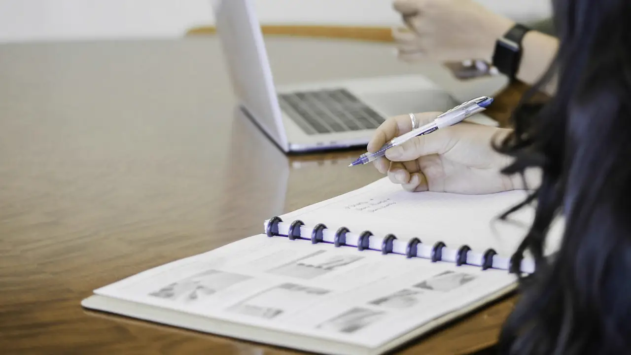 A person writing in a notebook beside a laptop