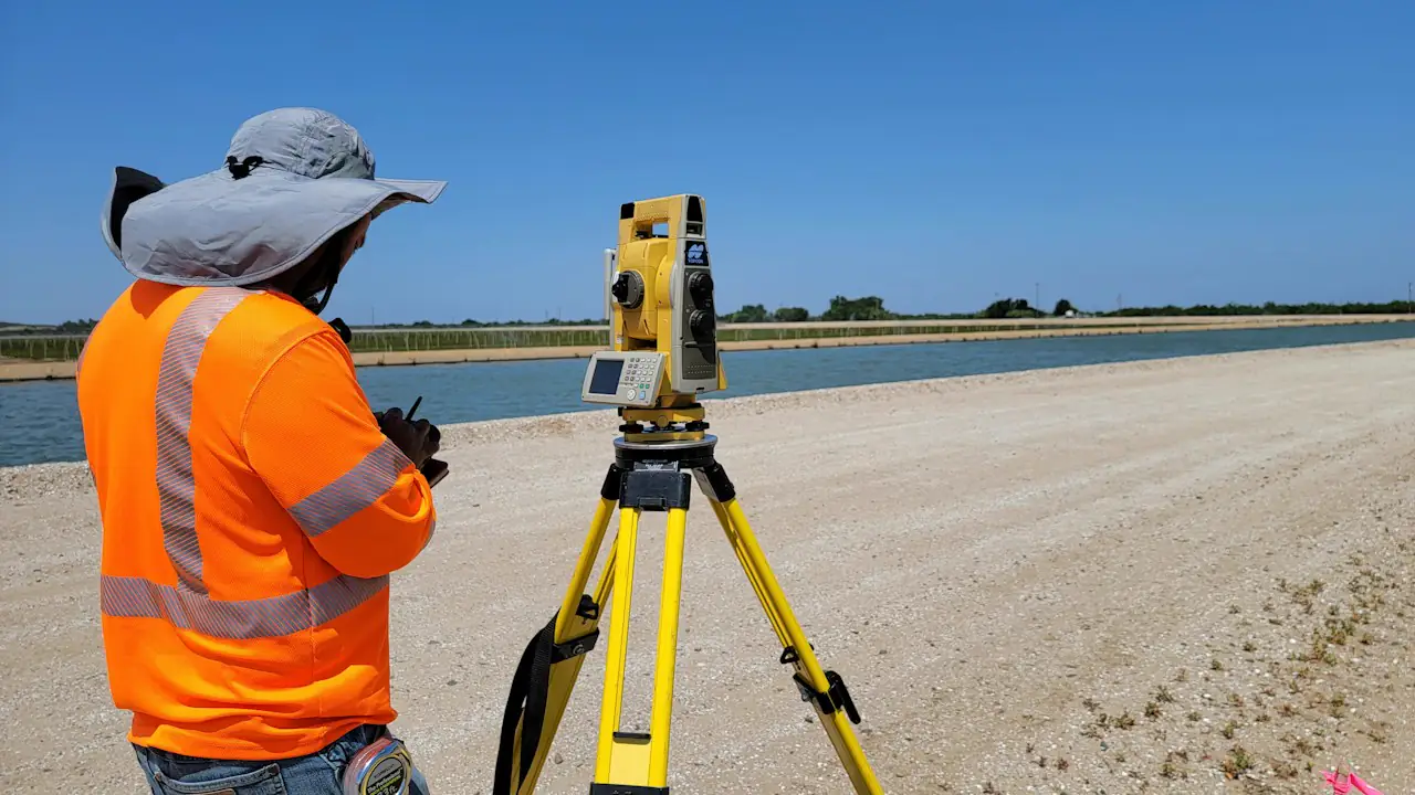 A surveyor in a high visibility vest and sun hat working with a total station on a gravel site